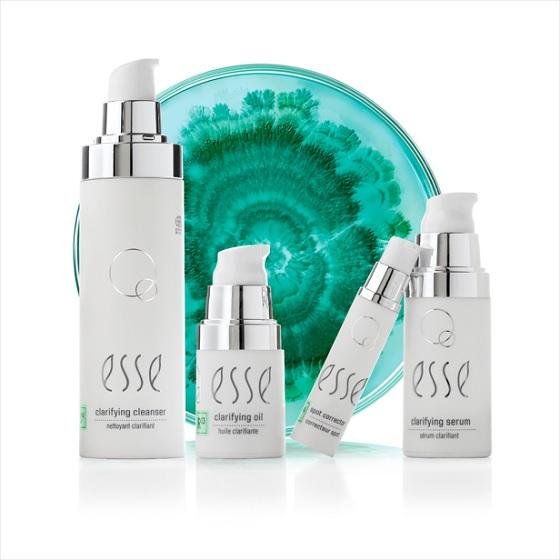 Four white bottles of Esse skincare products with silver pumps are arranged in front of a green and white decorative glass plate. The products are labeled clarifying cleanser, clarifying oil, and clarifying serum.