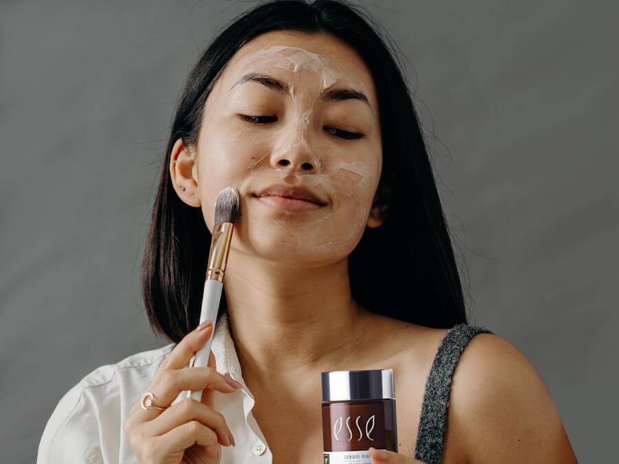 A woman gently applies a facial mask to her face with a brush, holding a jar labeled "Esse" in her other hand, against a neutral gray background.