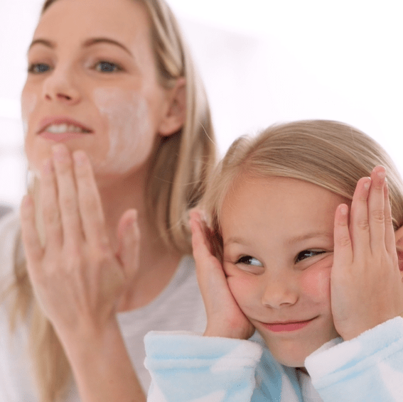 A woman applies cream to her face while a young girl next to her smiles and holds her hands to her cheeks, both wearing light-colored robes in a bright setting.