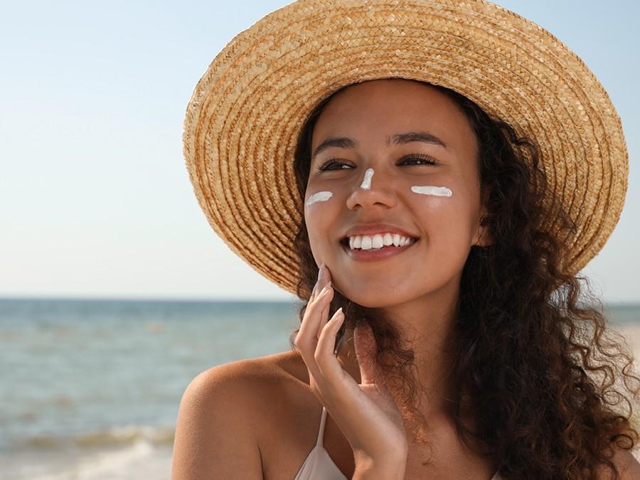 A smiling woman wearing a wide-brimmed straw hat stands on a beach with sunscreen applied to her cheeks and nose, touching her face and looking into the distance. The ocean and sky are in the background.