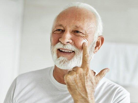 Smiling older man with white hair and beard, wearing a white shirt, applies cream to his cheek with one finger in a bright room.