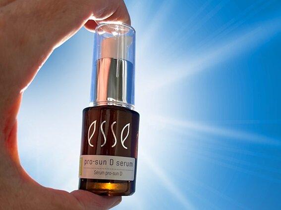 A hand holds up a small bottle of Esse Pro-Sun D Serum against a bright blue sky with sun rays shining in the background.