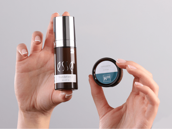 A person holds a bottle of Esse foundation in one hand and a small container of H2O concealer in the other against a light grey background.