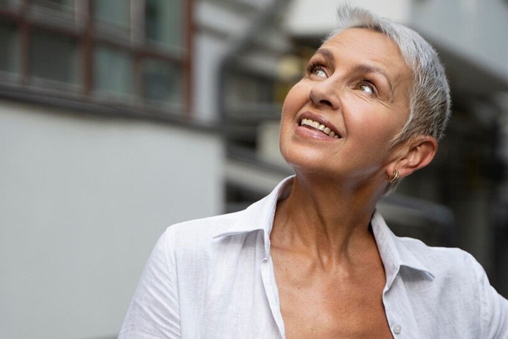 Smiling older woman with short gray hair wearing a white shirt looks up, standing outdoors in front of a blurred building background.
