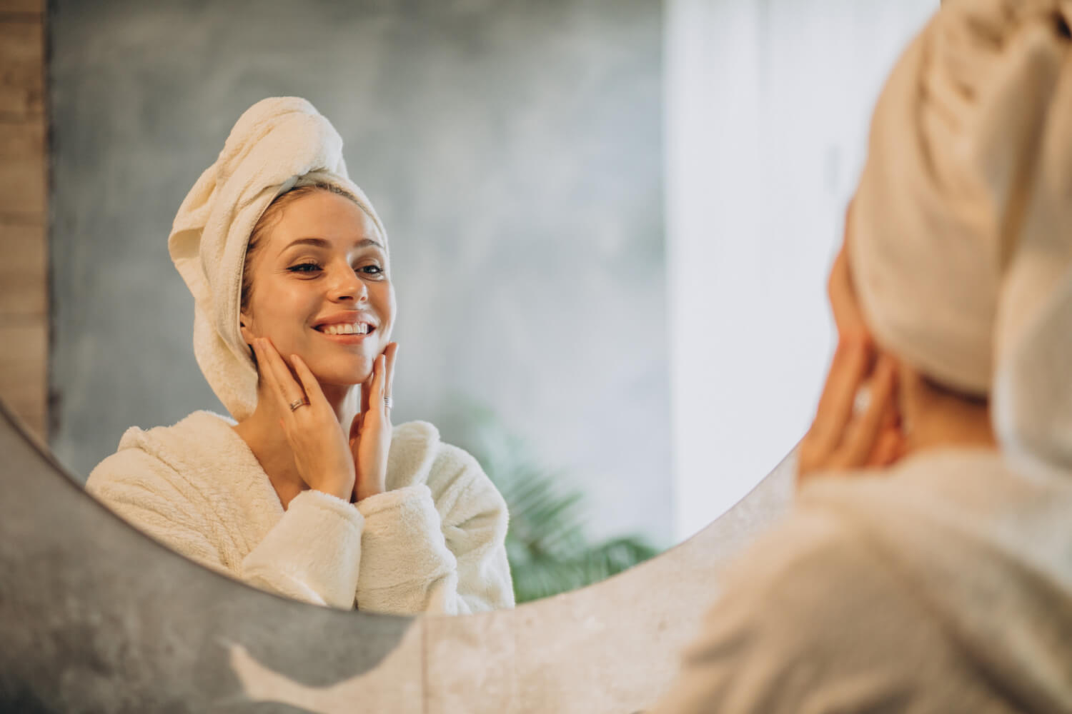 A woman with a towel wrapped around her head and wearing a bathrobe stands in front of a mirror, smiling and touching her face with both hands in a bright, cozy bathroom.