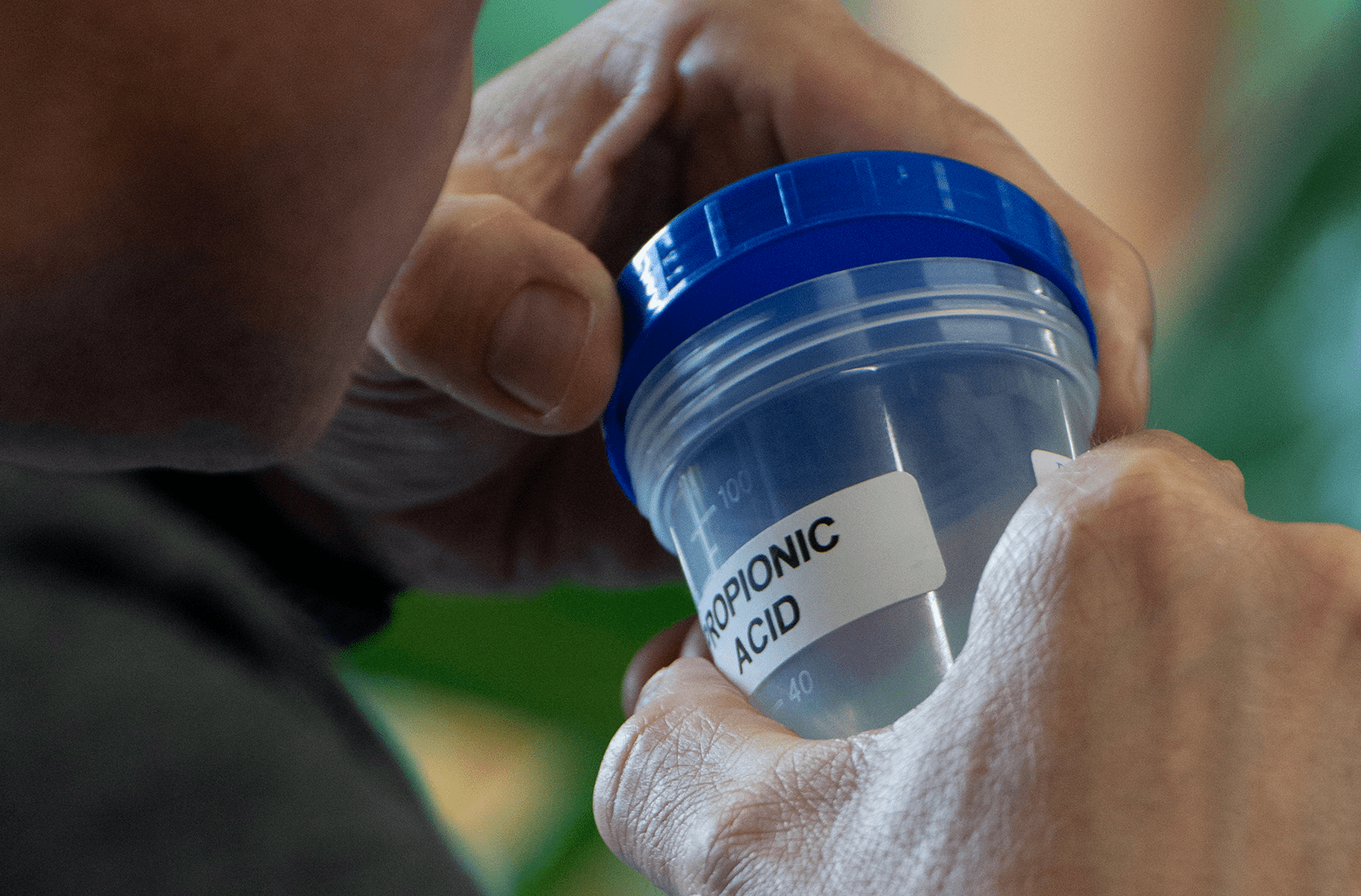 A person holding a clear plastic container with a blue lid, labeled "PROPIONIC ACID," positioned near their nose as if smelling the contents.