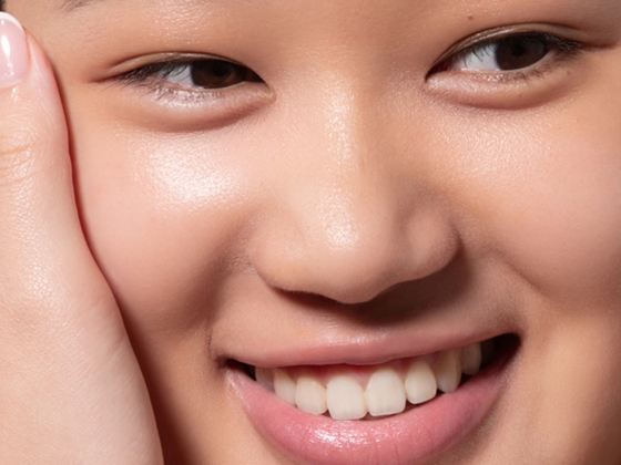 A close-up of a smiling person’s face with smooth, glowing skin. One hand gently touches their cheek, showing well-groomed nails. Their eyes are looking slightly to the side.