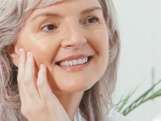 A smiling woman with light skin and gray hair gently touches her cheek, showing a healthy complexion. A plant is visible in the background.
