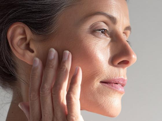 A close-up of a woman's face with smooth, healthy skin, gently touching her cheek with her fingers. She appears calm and is looking slightly upward in soft, natural light.