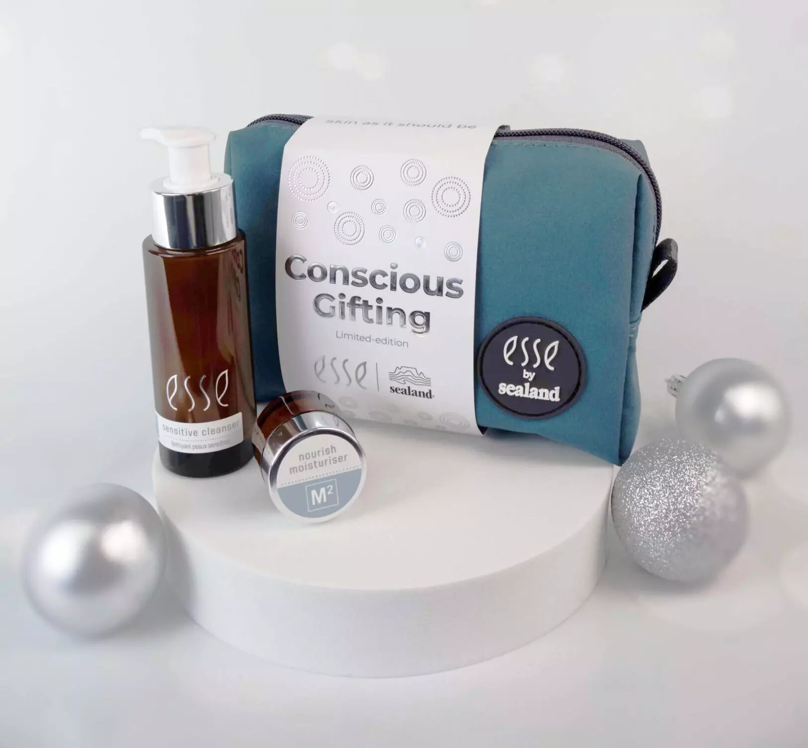 A blue toiletry bag labeled “esse by sealand” with a pump bottle, small moisturizer jar, and a card reading “Conscious Gifting Limited edition,” surrounded by silver ornaments on a white display. Ontdek het Esse Mist-assortiment.