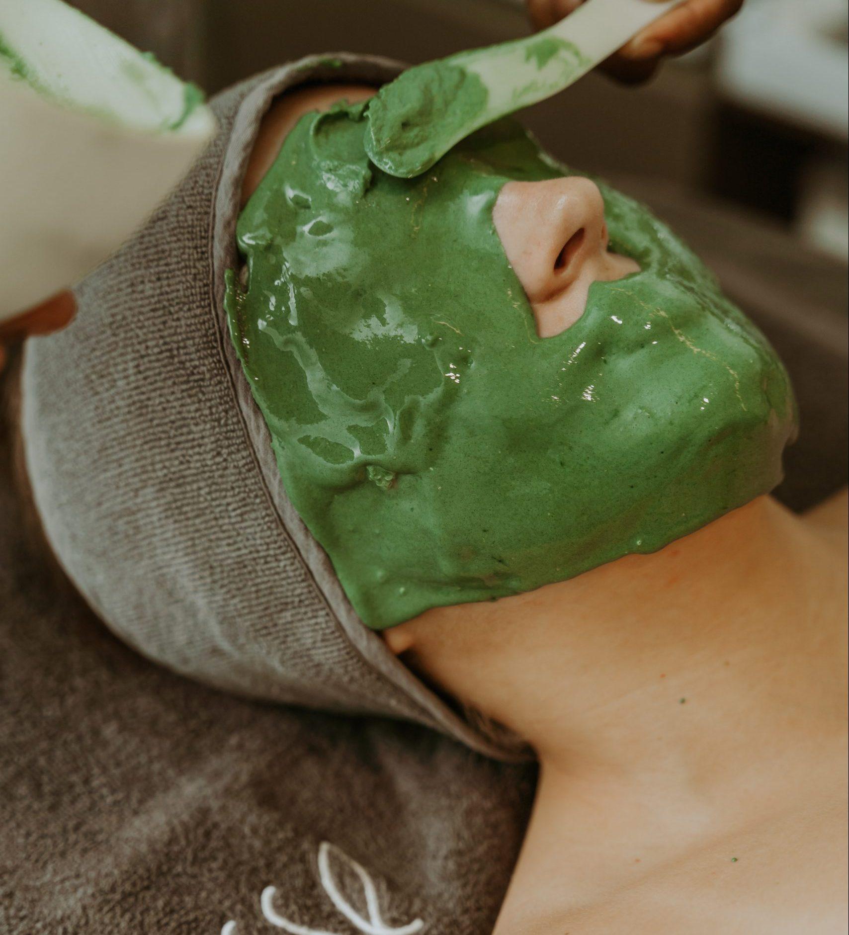 A person lies down with a thick green facial mask being applied to their face using a spatula. They are wearing a gray towel wrapped around their head, and their eyes are closed, relaxing during the spa treatment.