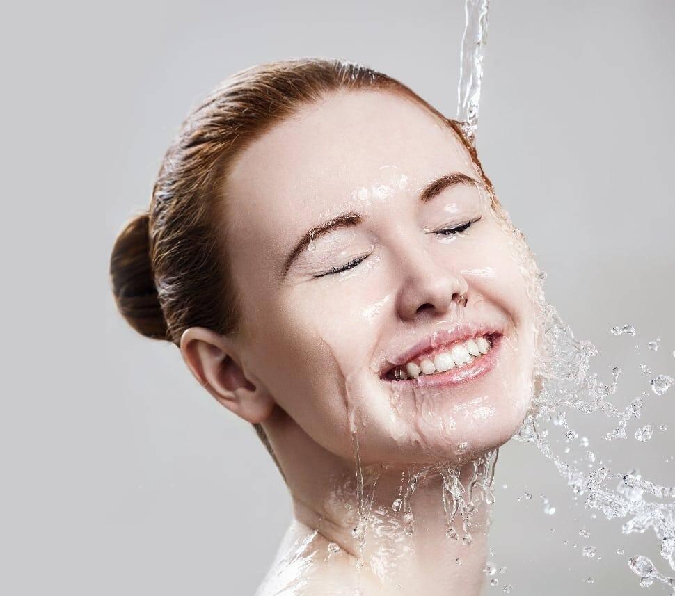 A woman with fair skin and brown hair in a bun smiles with her eyes closed as water splashes over her face. The background is plain and light-colored.