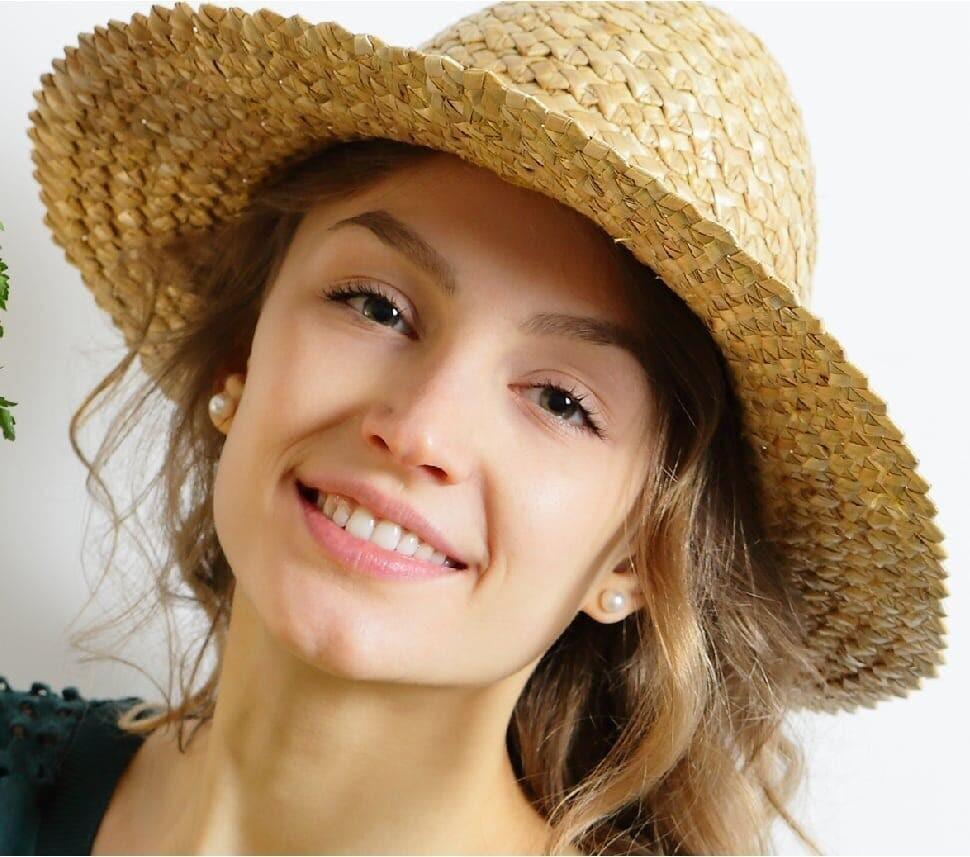 A young woman with wavy hair smiles at the camera, wearing a wide-brimmed straw hat and pearl earrings, against a plain white background.