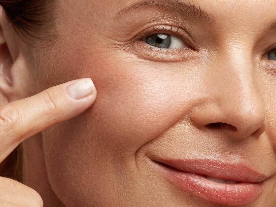 A close-up of a smiling person’s face, showing smooth skin and blue eyes. They are gently touching their cheek with one finger, highlighting healthy, radiant skin.