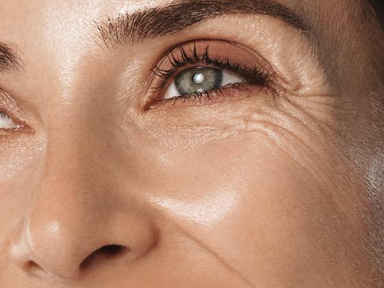 Close-up of a woman's face focusing on her eye area, showing natural skin texture and fine lines around the eye, with well-groomed eyebrow and smooth, glowing complexion.