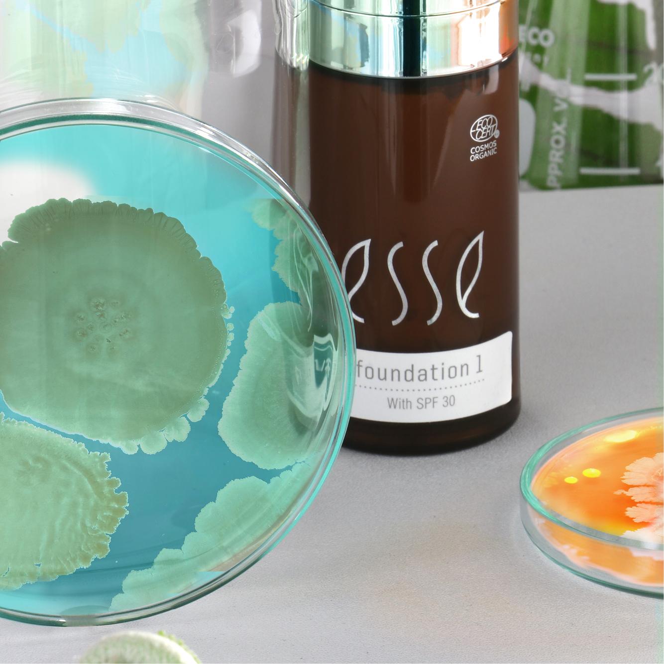 A bottle of foundation labeled "Esse Foundation 1 with SPF 30" is placed next to two petri dishes, one with teal bacterial cultures and another with orange and white cultures.