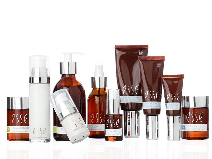 A selection of Esse skincare products in amber and white bottles and tubes of various sizes, grouped together against a white background. Some containers have pumps, droppers, or silver caps.