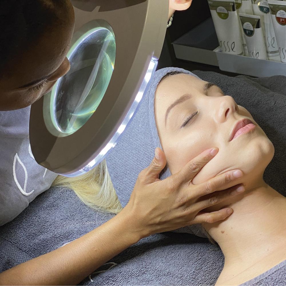 A person examines a woman's face under a magnifying lamp during a skincare treatment at a spa. The woman, eyes closed, lies on a towel-covered bed while the practitioner gently holds her chin. Skincare products are visible in the background.