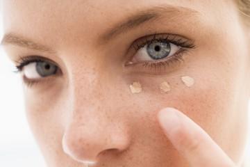 A close-up shows a person with blue eyes applying three dots of Esse Concealer under their eye and blending it with a finger, highlighting the eye area and makeup application.