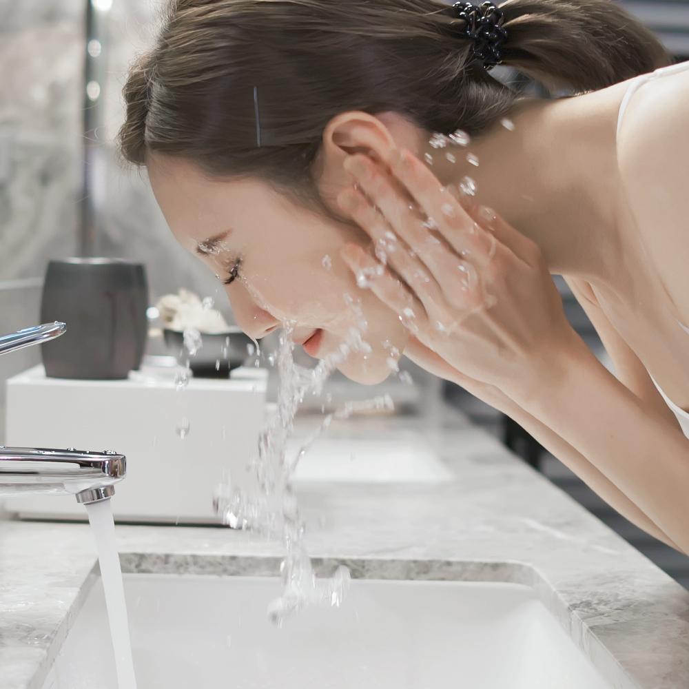 A woman with dark hair tied back washes her face with water from a sink faucet in a modern bathroom, splashing water onto her face with both hands.