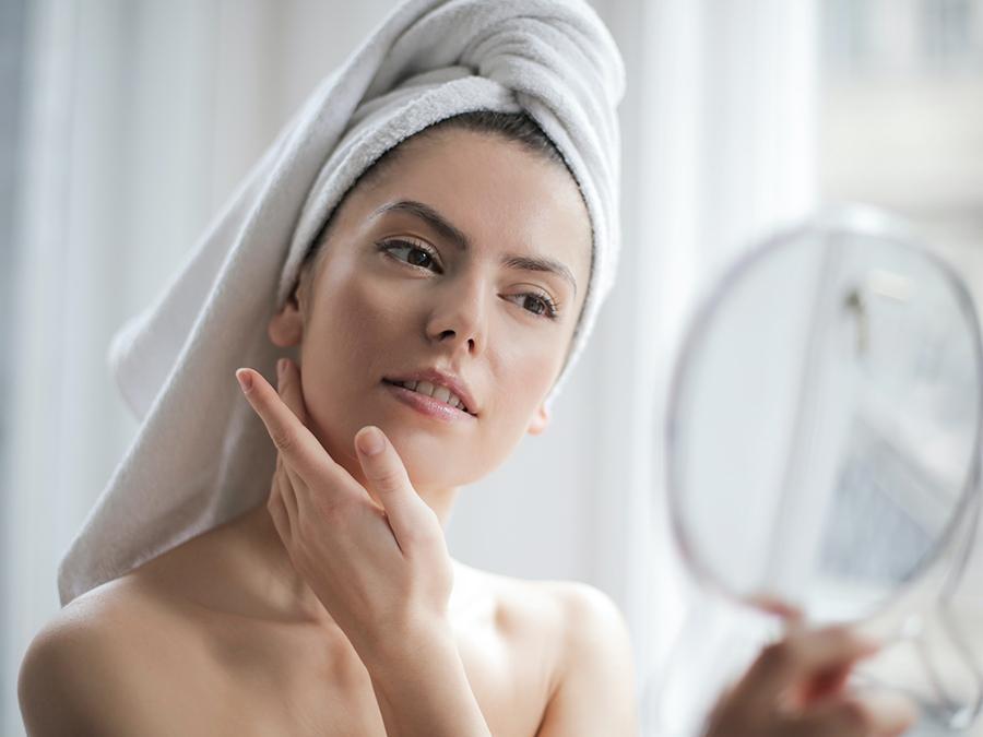 A woman with a towel wrapped around her head looks at herself in a handheld mirror while gently touching her face, appearing to examine her skin by a window with natural light.