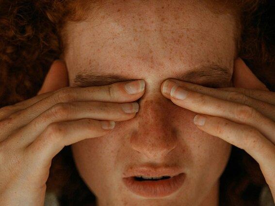 A person with freckles and curly hair is rubbing their eyes with both hands, appearing tired or stressed. Their eyes are closed and their facial expression suggests discomfort.