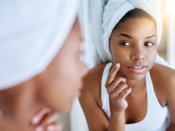 A woman with a towel wrapped around her head looks at herself in the mirror, gently touching her cheek with her finger, appearing thoughtful and focused on her skin.