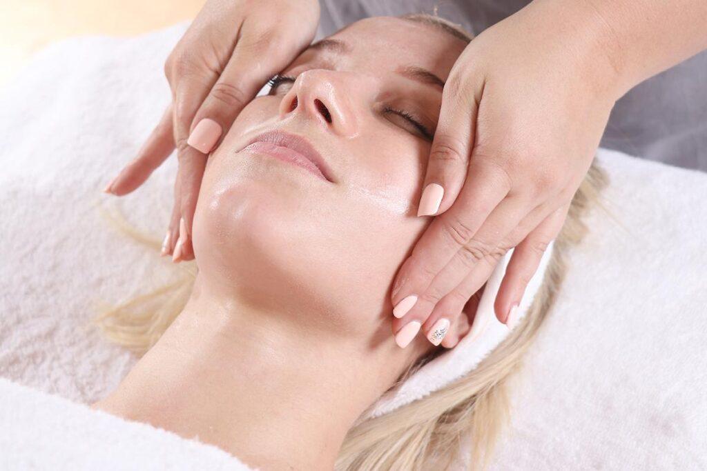A person with closed eyes lying on a towel receives a gentle facial massage from someone with manicured nails, creating a relaxing spa environment.