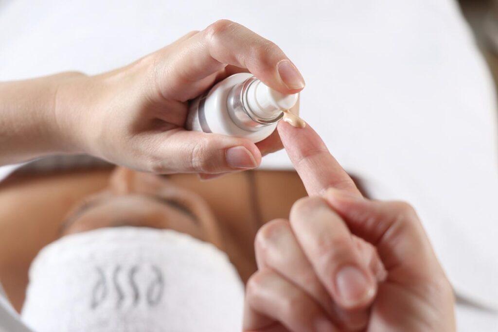 Close-up of a person dispensing a small amount of cream or serum onto their fingertip from a bottle, with another person lying down in the background wearing a headband and towel.