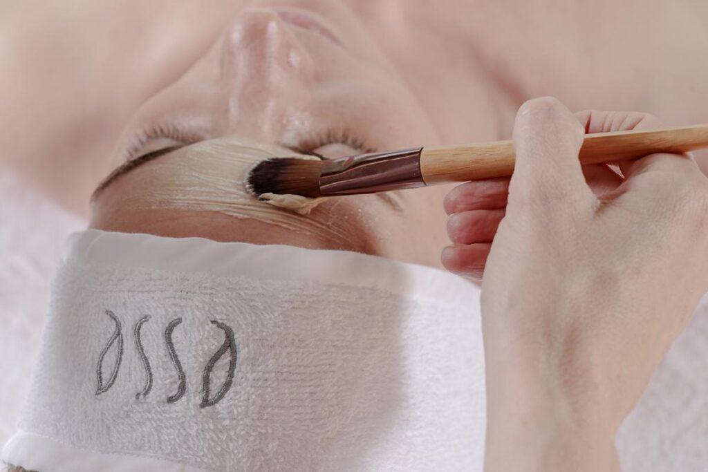 A person lying down with a white towel headband while another hand applies a facial mask with a brush to their forehead.