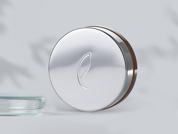 A round, silver metallic cosmetic container with a simple embossed leaf design on the lid sits upright on a light gray surface. A clear glass object is partially visible on the left. Shadows of leaves are seen in the background.