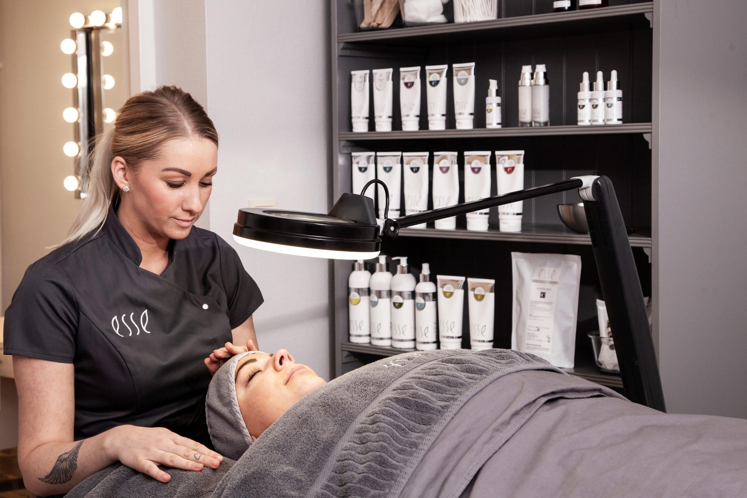 A spa therapist in a gray uniform prepares to give a facial treatment to a client lying on a treatment bed, covered with a gray blanket and towel, next to shelves stocked with skincare products.