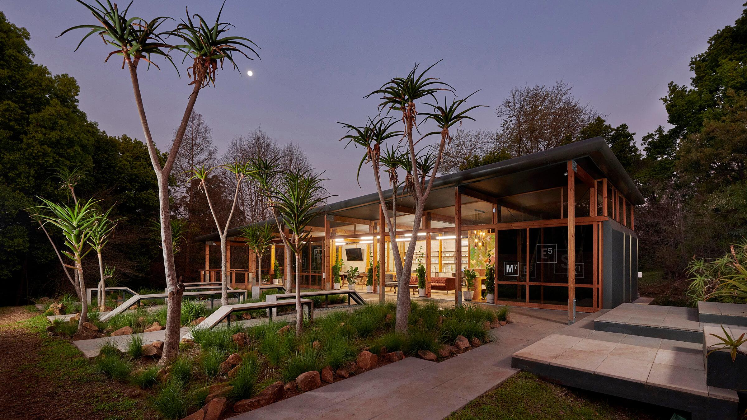 A modern glass house with wood accents, warmly lit inside, sits among landscaped gardens with path, tall spiky trees, and a fountain feature at dusk under a twilight sky with the moon visible.