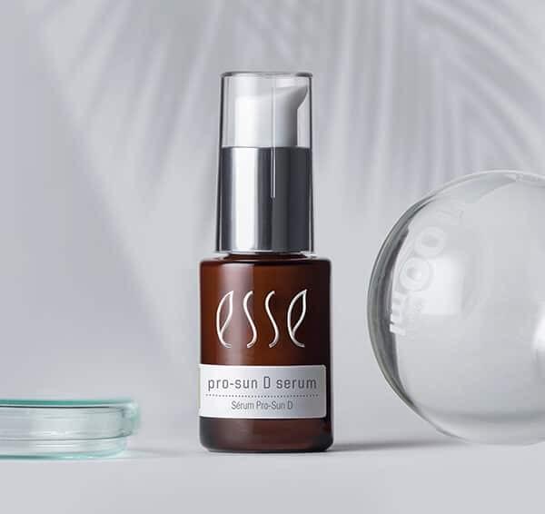 A brown glass bottle labeled "esse pro-sun D serum" with a silver pump cap stands next to a clear glass dish and a round glass object on a light gray background with palm leaf shadows.