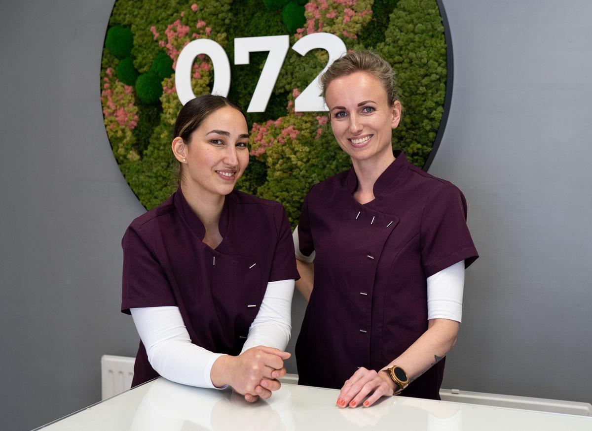 Two women wearing matching purple uniforms stand and smile at a reception desk. Behind them is a circular sign with green foliage and the numbers "072" displayed prominently.