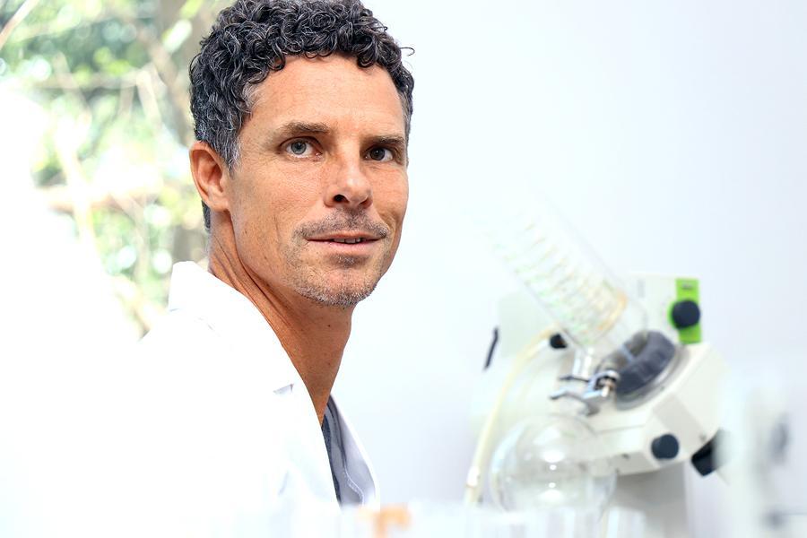 A man with short curly hair and a light beard, wearing a white lab coat, stands in a bright laboratory next to scientific equipment, looking towards the camera.