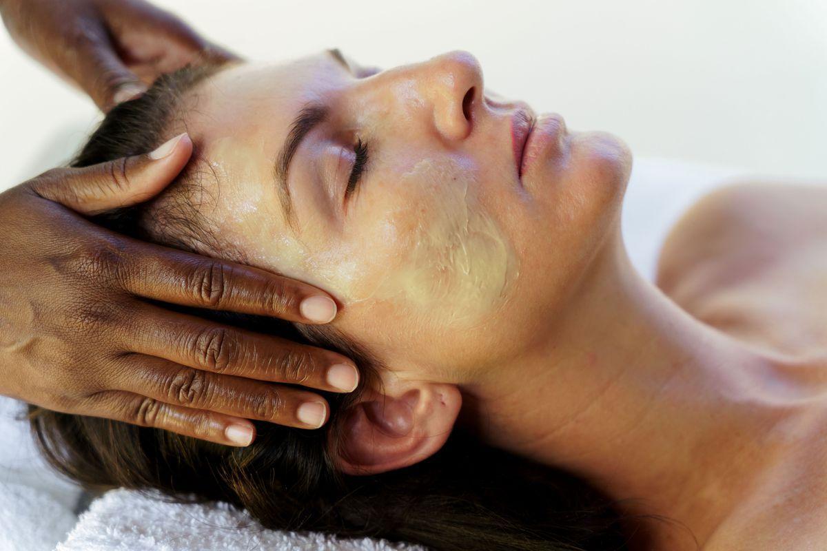 A person with their eyes closed receives a facial massage, with cream applied to their face and hands gently massaging their temples. The setting appears calm and relaxing.