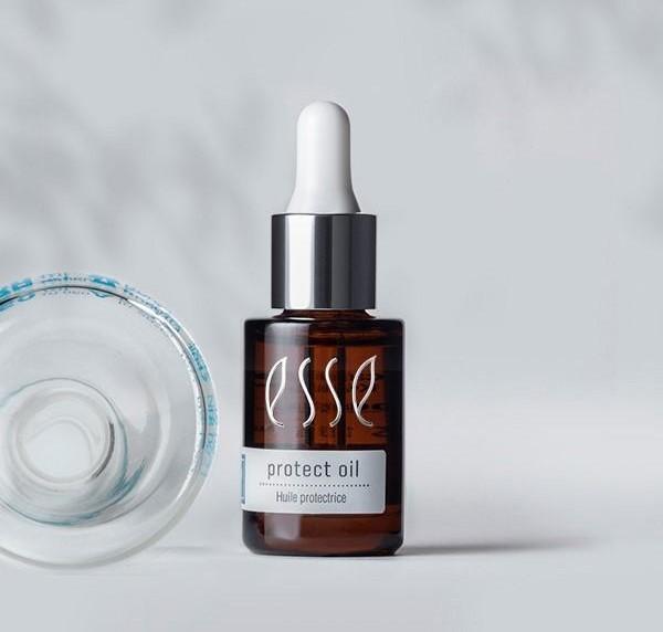A brown glass dropper bottle labeled "esse protect oil" stands upright next to a clear, round glass dish on a light surface with a soft, neutral background.